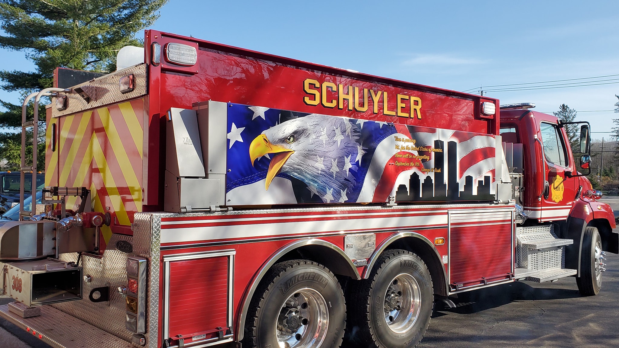 A red fire truck labeled “SCHUYLER” features a mural of an eagle, the American flag, and a city skyline on its side. The truck is parked outdoors near trees and a few buildings.