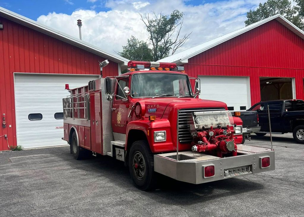 A bright red fire truck is parked in front of a red building with two large white garage doors on a clear day. A black pickup truck is parked beside the fire truck. Trees and blue sky are visible in the background.