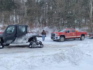 A person walks on a snowy road between a black off-road vehicle with snow tracks and a red fire department pickup truck, surrounded by snow-covered trees and hillside.