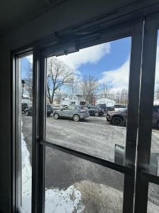 View through glass doors showing a snowy parking lot with several parked cars, leafless trees, and a white house in the background under a partly cloudy sky.