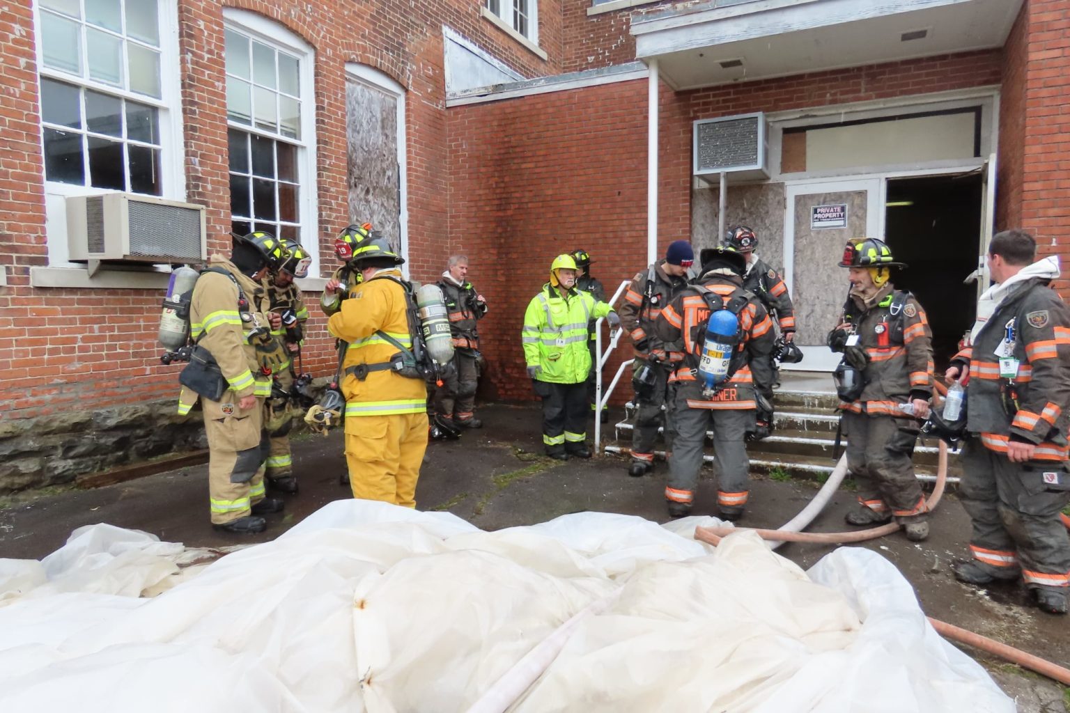 A group of firefighters in full gear gather outside a brick building with boarded windows. Some are holding hoses, while others appear to be in discussion near a large white tarp on the ground.