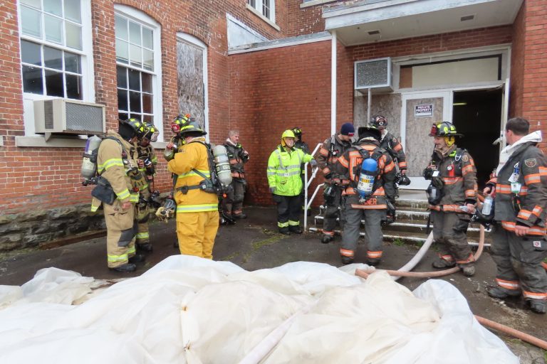 A group of firefighters in full gear gather outside a brick building with boarded windows. Some are holding hoses, while others appear to be in discussion near a large white tarp on the ground.